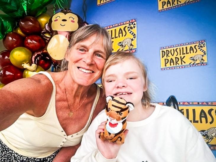 A woman and a young girl smile together at Drusillas Park, East Sussex, at a Momentum Children's Charity Dreamnight event. The girl holds a tiger soft toy, with a colourful balloon arch behind them.