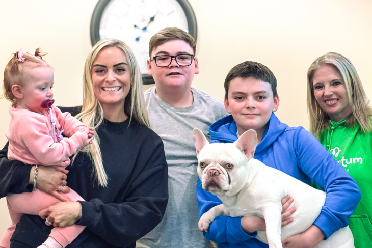 A smiling family of four, supported by Momentum Children's Charity, pose together indoors. A woman holds a baby girl dressed in pink, while two boys stand beside them, one cradling a white French Bulldog. A Momentum Family Support Worker in a green Momentum hoodie stands smiling with the family.