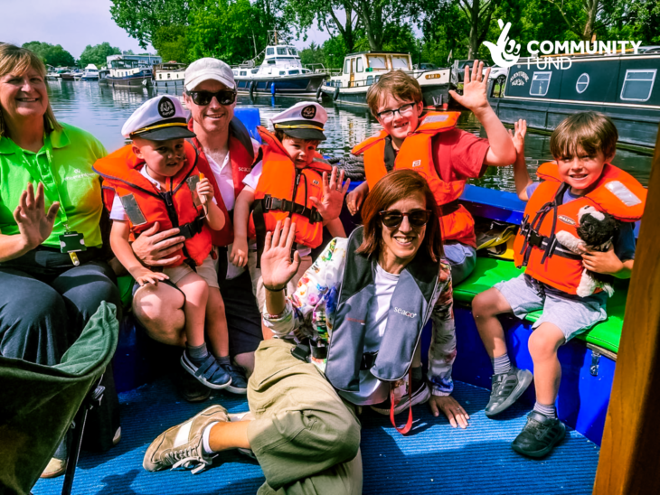 A supported family sit at the front of the boat waving at the camera. A Momentum family support worker sits beside them.